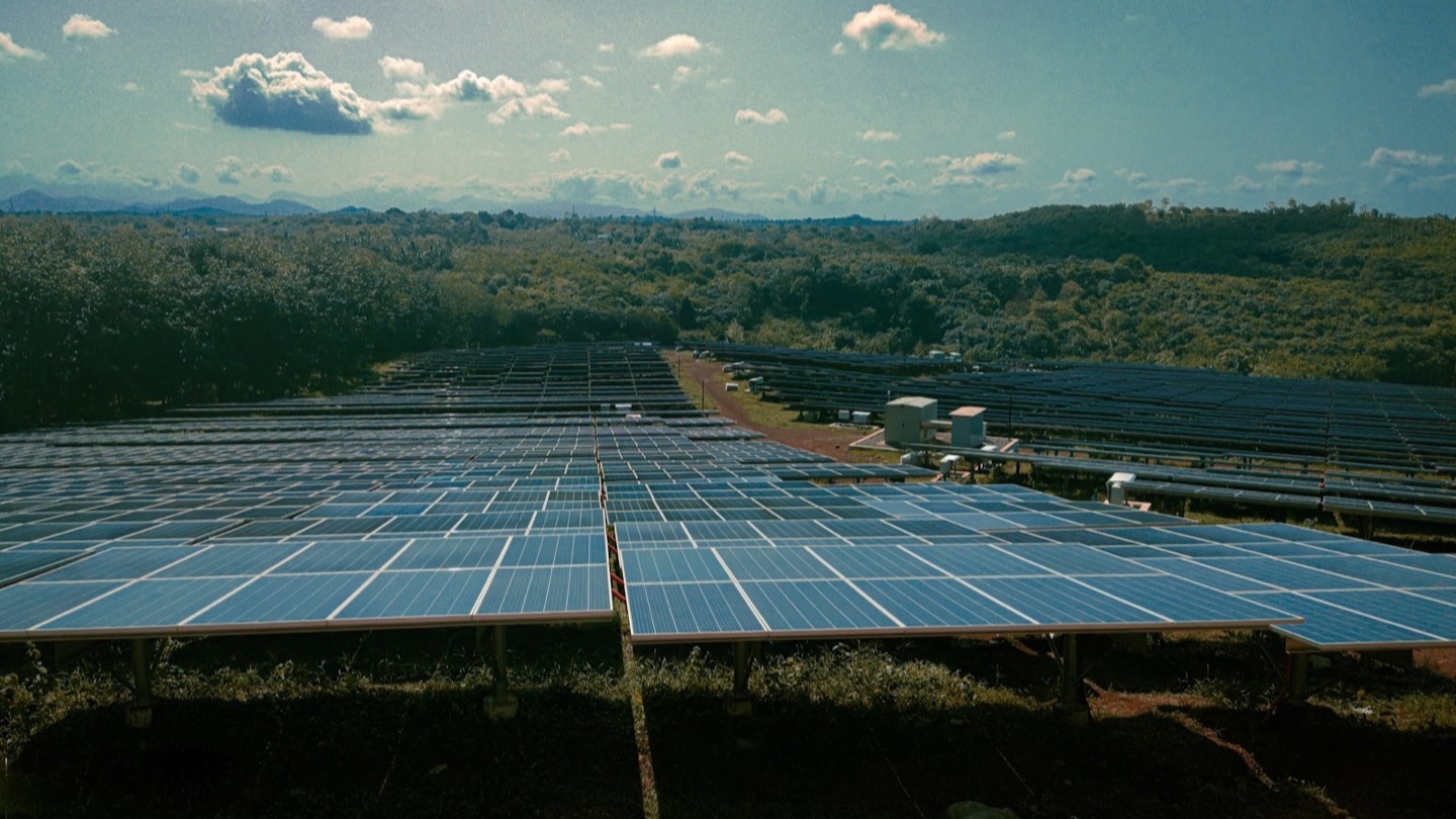 A large solar farm with many rows of panels set in a green, hilly landscape